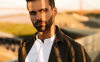 A man with dark hair and a beard stands outdoors, wearing a white shirt and a dark jacket. Sunlight casts a shadow across his face. The background is blurred, showing an outdoor walkway and distant structures.