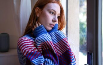 A young woman with long red hair and a colorful, striped sweater stands by a window, gazing outside with a thoughtful, pensive expression, her hands gently touching her neck.