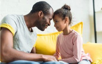 A man and a young girl sit on a yellow couch, touching foreheads and smiling warmly at each other, showing a close and affectionate bond.