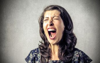 A woman with long dark hair, wearing a patterned top, is standing against a gray background. Her eyes are closed and her mouth is wide open, as if she is screaming or shouting.