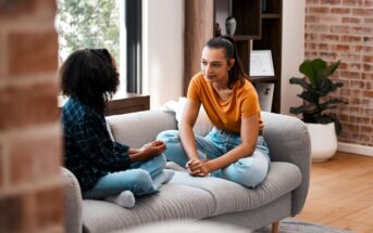 Two young women sit on a gray couch in a cozy living room, facing each other and having a serious conversation. One wears an orange shirt and jeans, the other a plaid shirt and jeans. Natural light fills the space.