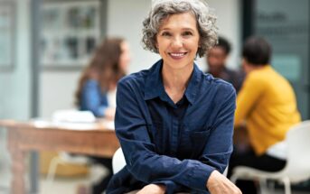 Smiling older woman with short curly gray hair wearing a blue shirt sits indoors with her arms crossed. In the blurred background, three people are talking around a table.