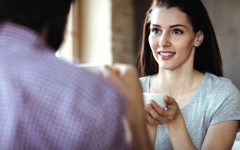 A woman with long brown hair smiles and holds a white coffee cup while sitting across from someone in a plaid shirt, engaging in conversation at a café.