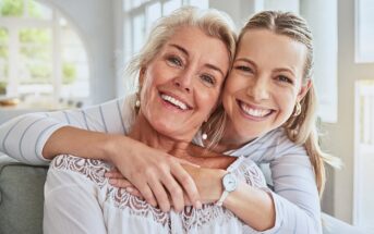 Two women sitting close together on a couch, smiling warmly at the camera. The younger woman hugs the older woman from behind. Both have light hair and are wearing light-colored, casual clothing in a bright, airy room.