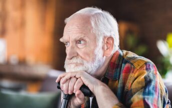 An elderly man with white hair and a beard is resting his hands and chin on a cane, looking thoughtfully into the distance. He is wearing a colorful plaid shirt and sitting indoors with a softly blurred background.