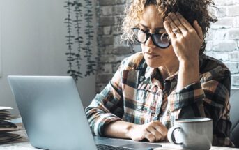 A woman wearing glasses and a plaid shirt sits at a desk with a laptop, holding her head in one hand and looking stressed. A coffee mug and paperwork are on the table next to her.