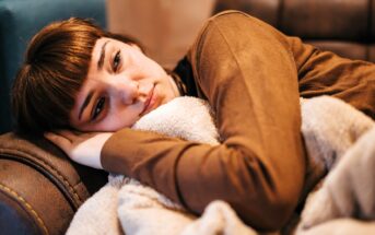 A woman with brown hair and bangs lies on a couch, resting her head on a pillow and hugging a soft beige blanket, looking tired or sad.