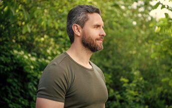 A man with short brown hair and a beard, wearing a green t-shirt, stands outdoors in front of lush green foliage, looking to the right with a calm expression.