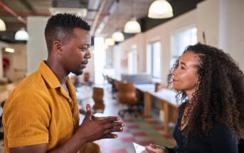 A man in a yellow shirt and a woman with curly hair are having a conversation in a modern, open-plan office. The woman is holding a smartphone and they appear engaged in discussion.