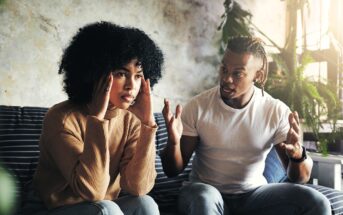 A woman sits on a couch touching her temples, looking frustrated, while a man next to her gestures with his hands and appears to be speaking intensely. They are indoors surrounded by plants.