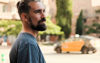 A bearded man with dark hair and a gray t-shirt stands outdoors, looking to the side. In the background, a yellow and black taxi passes by on a tree-lined city street.
