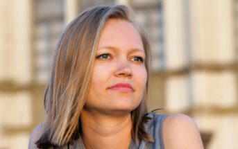 A woman with straight, shoulder-length blonde hair and blue eyes gazes thoughtfully into the distance, wearing a sleeveless gray top. The blurred background features a light-colored building.