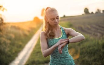 A woman in athletic wear stands on a rural dirt path at sunset, looking down and checking her smartwatch, with fields and soft sunlight in the background.