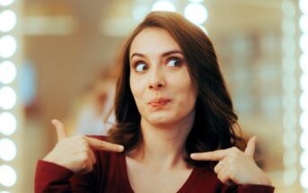 A woman with brown hair wearing a maroon top points at herself with both index fingers and makes a playful, surprised expression. The background is softly blurred with bright lights.