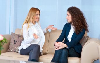 Two women sit on a beige sofa having a conversation. One woman gestures with her hands while speaking, and the other woman listens attentively with her hands resting on her knee. Both are dressed in business attire.
