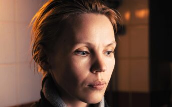 A person with short, light brown hair and light skin looks downward with a serious expression. Warm lighting illuminates their face, while a tiled wall is visible in the softly blurred background.