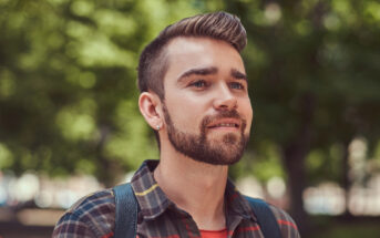 A young man with a beard and short hair, wearing a plaid shirt and backpack, stands outdoors with green trees blurred in the background. He is looking slightly upward and smiling softly.