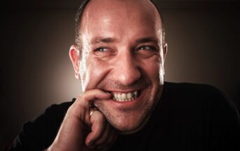 A man with light skin smiles playfully, biting his index finger. He is looking up and to the side, with a mischievous expression. The background is dark, and he is wearing a black shirt.