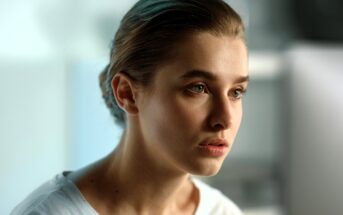 A young woman with light skin and brown hair pulled back looks thoughtfully ahead. She wears a white shirt and is in a softly lit, blurred indoor setting.