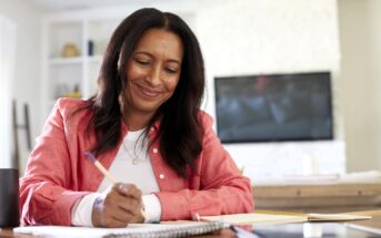 A woman with long dark hair and a pink shirt sits at a table, smiling as she writes in a notebook. A coffee mug, tablet, and books are on the table, with a blurred TV and shelves in the background.