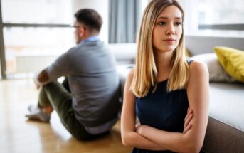 A young woman with crossed arms looks away, appearing upset, while a man sits on the floor with his back to her. They are indoors in a bright living room, suggesting tension or disagreement.