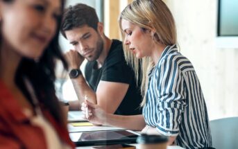 Two people sit at a table working together, with a tablet, notebooks, and coffee cups in front of them. One woman writes on the tablet while a man watches. Another person is blurred in the foreground.
