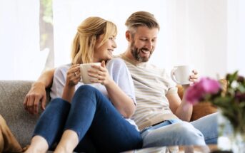 A smiling couple sits close together on a couch, each holding a mug. They appear relaxed and happy, looking at each other, with natural light coming from a window behind them and flowers blurred in the foreground.