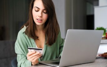 A woman with long brown hair sits at a table using a laptop and holding a credit card, appearing focused as she looks at the card. She is wearing a green shirt in a modern indoor setting.