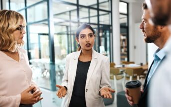 Four people stand in an office, engaged in conversation. One woman in a white blazer speaks while gesturing with her hands. Two others listen intently, and one person holds a coffee cup. Glass walls and tables are visible in the background.