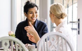 Two women sit at a table near a window, having a friendly conversation. One woman, with dark hair in a bun, smiles warmly at the other, who has blonde hair in a low bun. A cup and a vase with a rose are on the table.