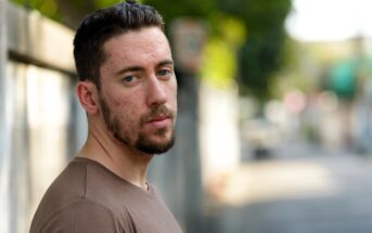 A young man with short dark hair and a trimmed beard stands outdoors, wearing a brown shirt. He looks at the camera with a neutral expression. The background is blurred, showing a street and greenery.