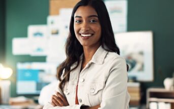 A woman with long dark hair and a light-colored jacket smiles confidently with her arms crossed, standing in an office with a dark green wall and blurred bulletin board in the background.