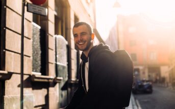 A smiling man with a backpack stands on a sunlit city street, wearing a black hoodie. Warm sunlight creates a bright glow and long shadows on the buildings and street in the background.