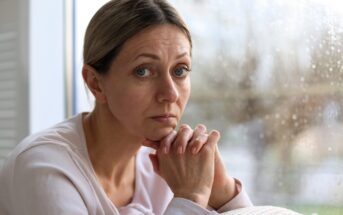 A woman with light brown hair pulled back rests her chin on her hands and looks thoughtfully at the camera. She sits by a window with raindrops visible on the glass, creating a somber atmosphere.