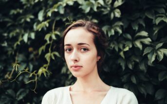 A young woman with light skin and brown hair stands in front of a wall covered in green leafy plants, looking directly at the camera with a neutral expression. She is wearing a white top.