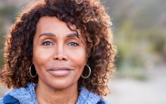 A middle-aged woman with curly hair and hoop earrings looks calmly at the camera outdoors, wearing a blue sweater. The background is softly blurred with natural greenery.