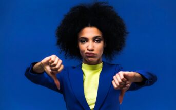 A woman with curly hair, wearing a blue blazer and yellow top, stands against a blue background, looking serious and giving two thumbs down.