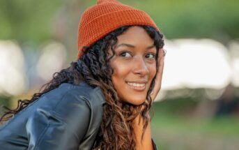 A young woman with curly hair, wearing an orange knit beanie and a black jacket, smiles while resting her face on her hand outdoors with a blurred green background.