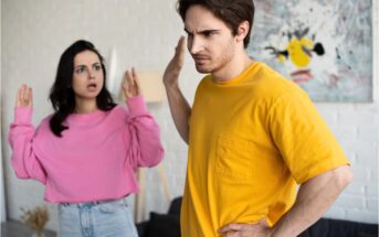 A woman in a pink sweater gestures with raised hands, looking upset, while a man in a yellow shirt turns away with a stern expression, raising his hand as if to stop her.