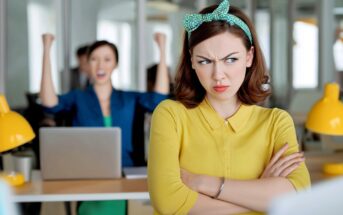 A woman in a yellow shirt stands with arms crossed, looking annoyed, while another woman in the background celebrates with raised fists at a desk in an office setting.