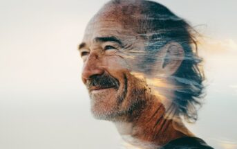 A smiling older man with gray hair and a beard, overlaid with a double exposure of a scenic ocean sunset, blending his profile with waves and sky.