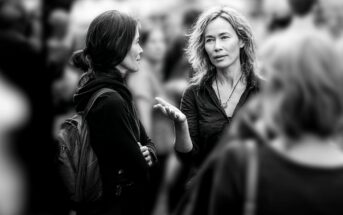 Two women stand facing each other in conversation outdoors, surrounded by a blurred crowd. One woman gestures with her hand while speaking; both are dressed casually. The image is in black and white.