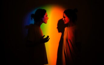 Two women face each other in a dark room, standing in front of a colorful rainbow light. One woman gestures and appears to be yelling, while the other looks upset. Both cast shadows on the wall behind them.