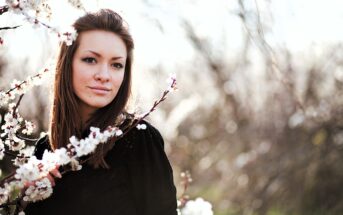 A woman with long brown hair stands near a branch of blooming flowers. She wears a black top and looks towards the camera with a gentle expression. The background is softly blurred, highlighting the bright blossoms.