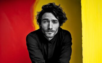 A man with wavy dark hair and a trimmed beard wears a black button-up shirt. He poses against a vibrant background divided between red and yellow, with an expressive and thoughtful look on his face.