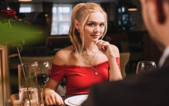 A woman with blonde hair, tied in a ponytail, sits at a restaurant table adorned with wine glasses, a red rose, and plates. She wears a red off-shoulder top and jewelry, and looks across the table with a soft smile, while a man, slightly blurred, faces her.