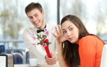 A man smiling and offering a bouquet of flowers to a woman who appears uninterested or uncomfortable. They are seated at a table in a bright setting with a window view. The woman is leaning back with a skeptical expression.