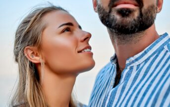 A woman with blonde hair and multiple ear piercings smiles and looks up at a man with a beard who is gazing into the distance. Both are wearing casual clothes and appear to be outdoors, with a blurred background suggesting they are near the water.