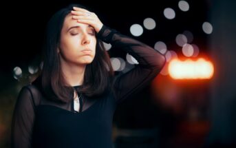 A woman with long dark hair stands with her eyes closed and her right hand on her forehead, appearing stressed or tired. She is wearing a black top and is set against a blurry background with bokeh lights.