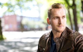 A man with short blonde hair and a trimmed beard is wearing a brown leather jacket and a navy shirt. He looks off into the distance with a thoughtful expression. The background is a blurred outdoor setting with trees and a building. Sunlight filters through the leaves.